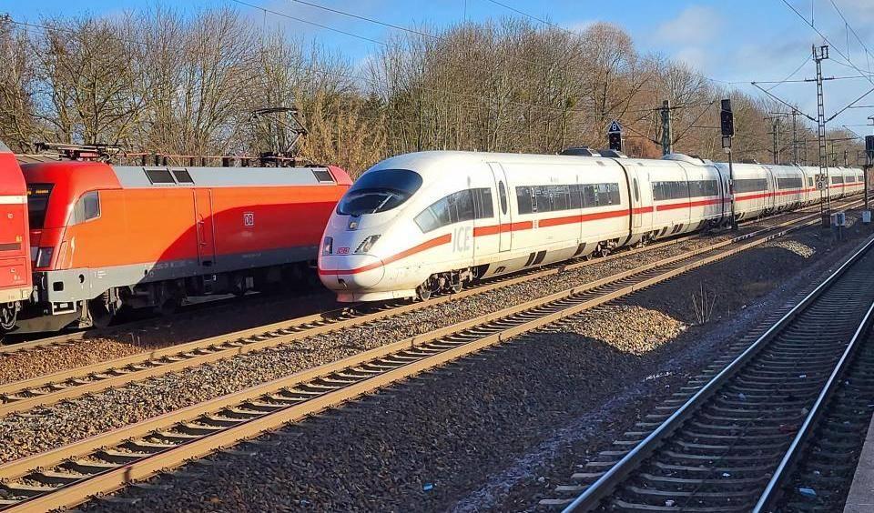 Ein ICE-Zug und eine rote Lokomotive auf nebeneinanderliegenden Gleisen vor kahlen Bäumen unter blauem Himmel